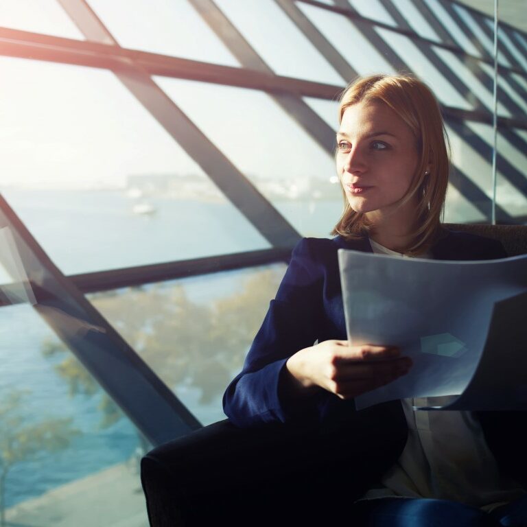 Portrait of young elegant woman sitting in modern office interior holding papers and pensively gazing out of the window, filtered image with flare sun light from the window